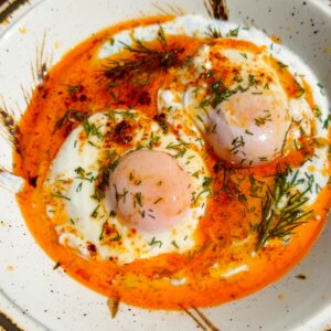 A plate of Turkish poached eggs (Çılbır) with creamy yogurt, runny eggs, and spiced butter, served with toasted crusty bread.