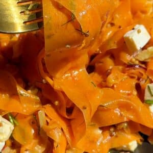 A fork holding a bite of carrot ribbon salad with feta, toasted walnuts, and fresh dill, coated in a zesty orange-honey dressing.