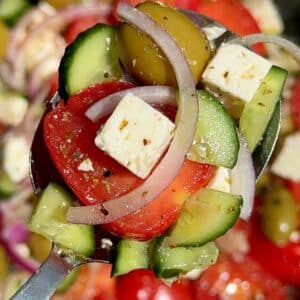 A close-up of a spoonful of Greek salad showing diced cucumber, cherry tomato halves, a cube of feta cheese, a slice of red onion, and a green olive, all coated lightly with dressing.