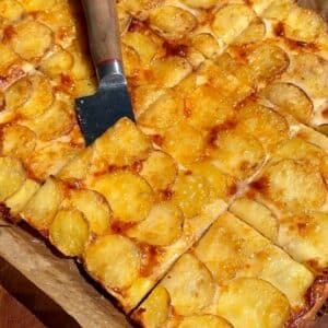 Slices of ham and potato pizza bake on a cutting board, showing melted cheese and golden potatoes, ready to serve.