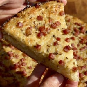 A slice of German Onion Pie (Zwiebelkuchen) held in hand, showing its golden crust, creamy onion filling, and bits of smoked bacon.