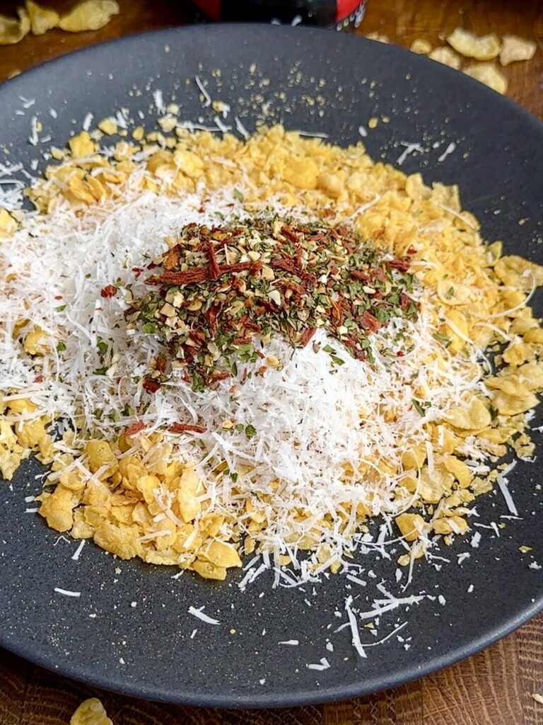 A shallow dish with crushed cornflakes, Italian herbs, and grated Parmesan mixed together, ready for coating the chicken.