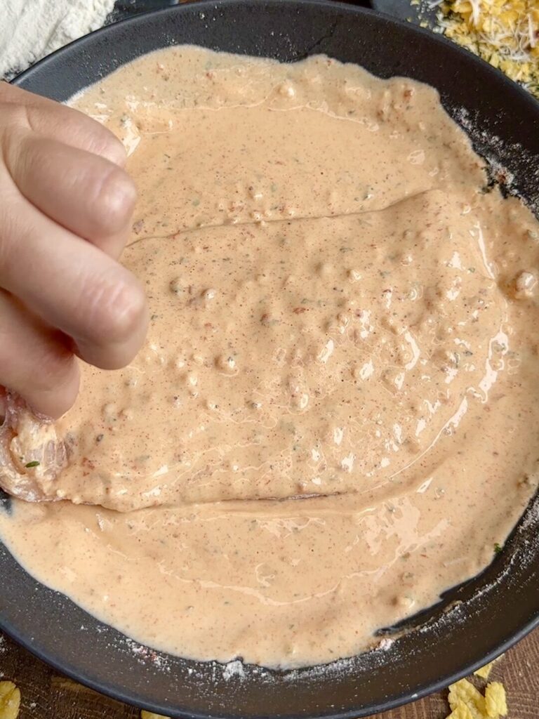 A chicken breast being dipped into the creamy yoghurt and spice mixture, fully coating it before adding the cornflake crust.