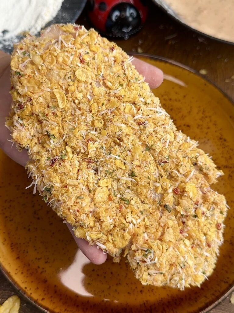 A chicken breast being pressed into the cornflake, Parmesan, and herb mixture, ensuring an even, crispy coating.