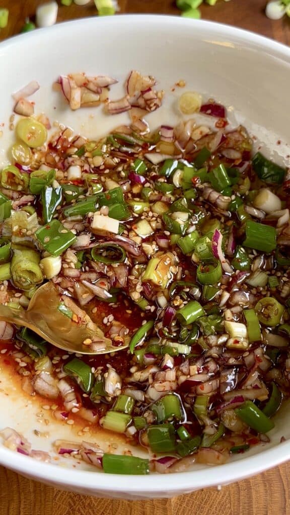 Mixing sesame oil, soy sauce, rice vinegar, agave syrup, chili sauce, chili flakes, red onion, garlic, spring onions, and sesame seeds to make a flavorful Asian-style marinade for cauliflower salad.